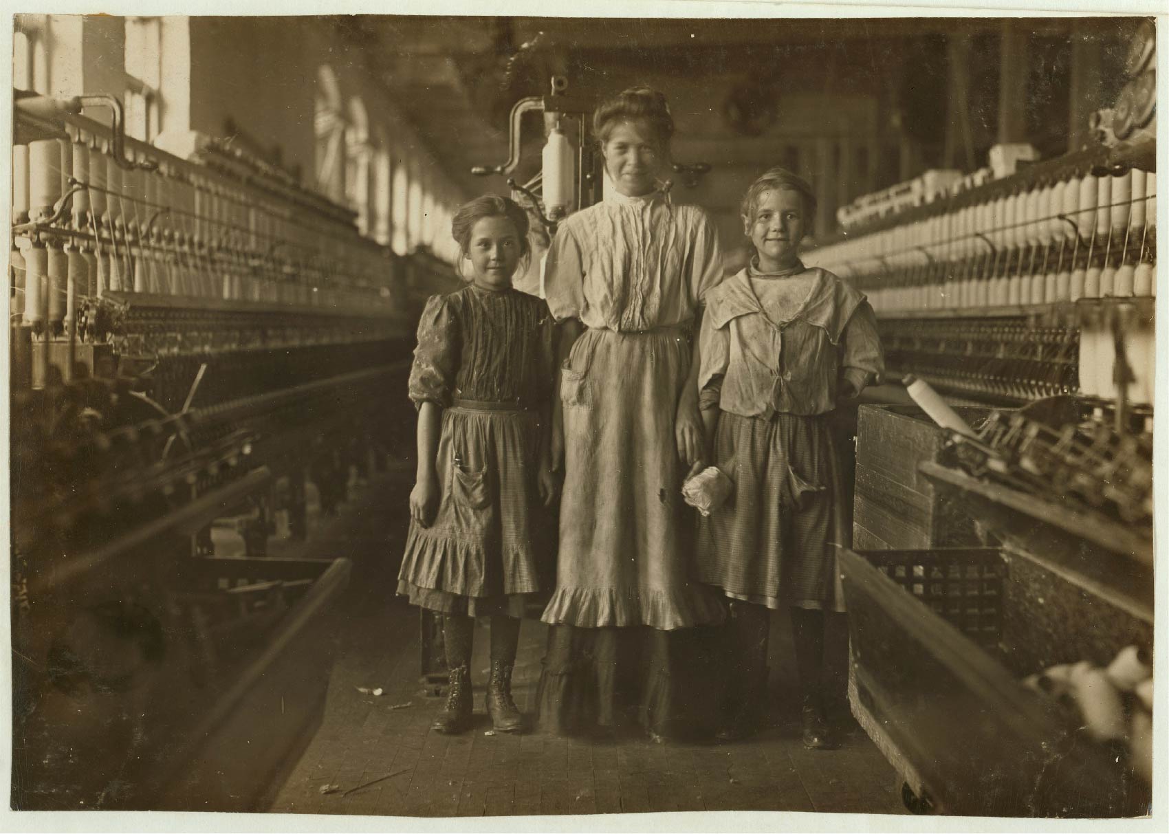 A vintage photograph of a woman flanked two girls standing in an  textile factory.